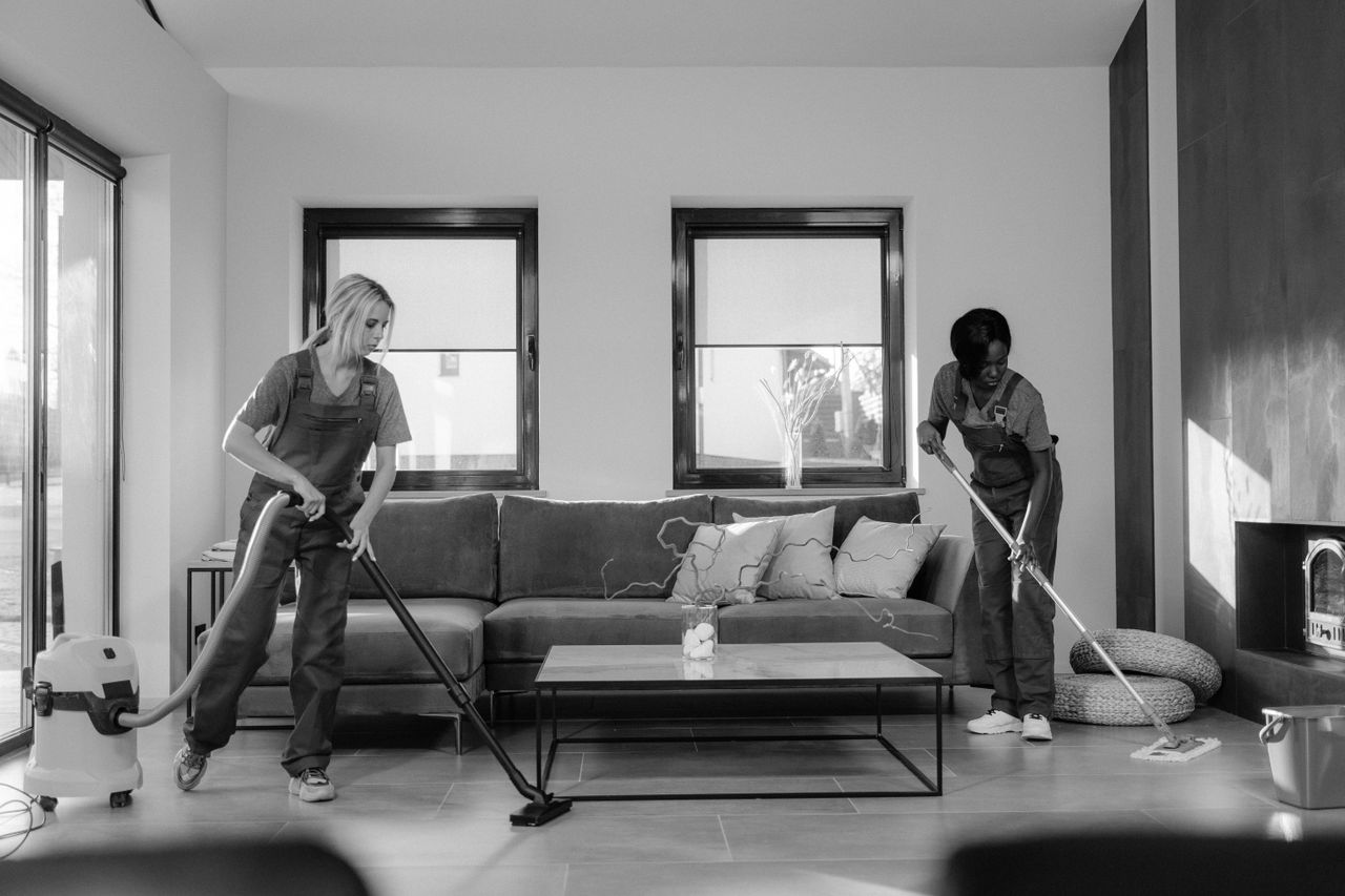 Two women dressed for work are tidying a contemporary living room with a vacuum cleaner and a mop.