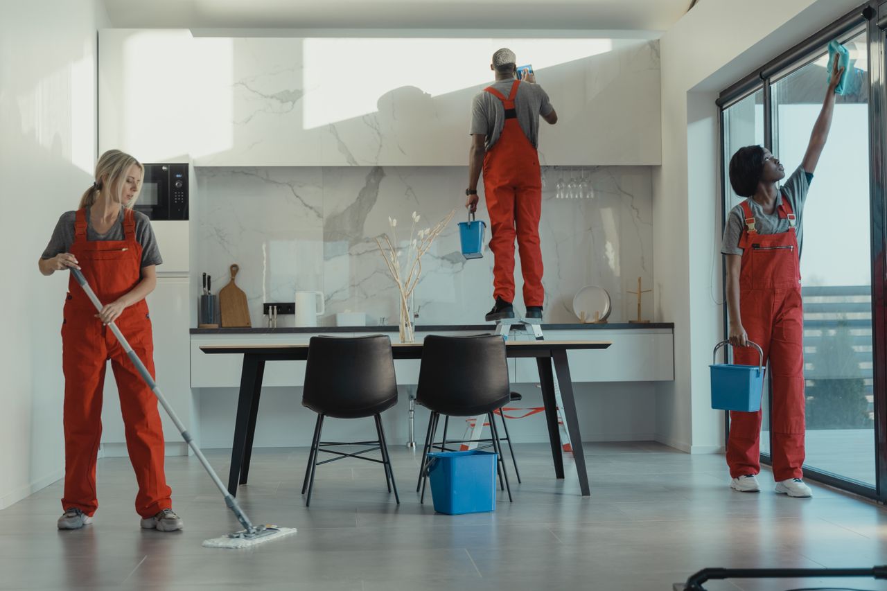Team of cleaners in red uniforms are mopping the floor and wiping glass in a contemporary dining area.